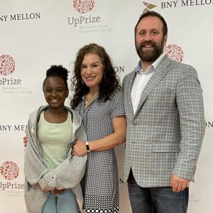Dr. Crystal Morrison, Meerkat Village co-founder and CEO, stands in the center in front of the UpPrize backdrop.  She stands with her 13 yr old daughter Zoe (on the left in the picture) and her husband Dr. Justin Bohn (on the right in the picture).  All a