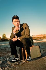 A portrait of Elvis impersonator, Victor Trevino, Jr. in character as the King of Rock and Roll. He sits upon a suitcase in a vintage 1950s style suite. His hair is styled in a 1950s pompadour, just like young Elvis wore.