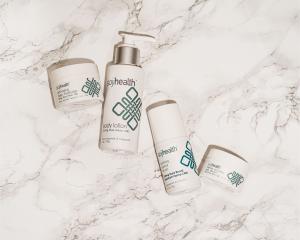 Soogee Health CBD skincare found in JCPenney Beauty includes Recovery Eye Cream, Anti-Aging Daily Moisturizer, Body Lotion, and Soothing Topical Rolls on All Lay Flat on a white marble surface.