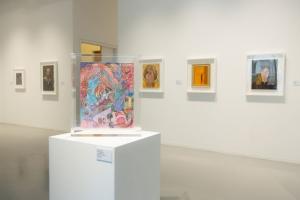 A photo of a large white room with several pieces of art hung on the walls, as well as a framed piece on a pedestal in the center.