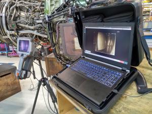 An aircraft engine suspended in a workshop with a camera-like device inserted into it. The camera feed is connected to a laptop, displaying what the camera sees on Aiir Innovations' software platform.
