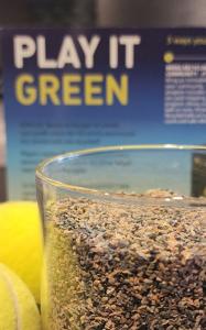 Bright yellow tennis balls sitting next to a canister of finely ground grey rubber, called Green Gold, in front of a blue and green poster that reads Play It Green.