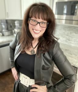 Nancy Manet, author of My Expansion, stands in her kitchen. She is wearing a leather jacket and belt and smiles nicely at the camera.