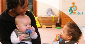 A caregiver sitting on the floor holding an infant with a toddler on the floor next to them
