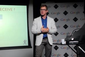 homem no palco com blazer cinza claro e calça azul com o logo do Google no fundo