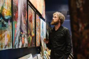 A bearded man looking at art paintings on the wall