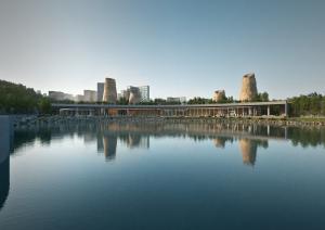 Image shows SOUR's proposal for the new Museum Library Cultural Complex from the lakeside view. In the back is a extended building with five rock chimneys rising up. In the front is a small lake and a lake boulevard. There is a blue sky and skyscapers in