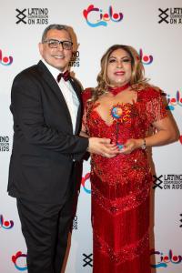 Elia Chinó standing to the right side of the image with blonde highlights in her dark hair, wearing a red dress with beaded work throughout. Elia Chinó is holding her award. Richard Izquierdo is on the left side of the image wearing a black suit with salt