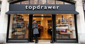 A photo of a man leaning against the doorway of Topdrawer, a store with bags, house shoes, vintage cameras, eyewear, and more.