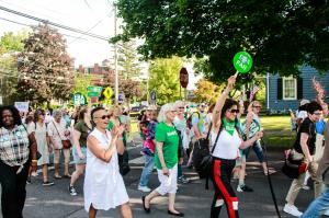 Dozens of women walk on a tree lined road in Seneca Falls, New York. Many are smiling. It is daytime in the summer, the sky is blue, and a few of the women are holding green round signs which say the "ERA Now" in white letters.