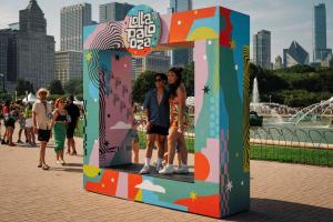 Two people standing inside a large square sculpture. The sculpture is wrapped in multi-colored Lollapalooza branding. In the background are the tall buildings of the Chicago skyline.