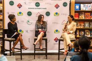 Horizontal photograph of three women sitting on stools speaking to an audience. In the foreground we see the heads of some people watching. At the bottom of the image there is a banner.