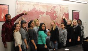 A group of students surround a red-toned map. Each points to where they're from, and many countries are represented.