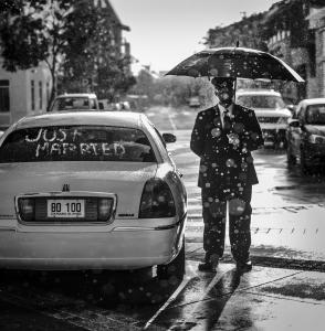 A chauffer stands next to his white car on a rainy day, he holds an umbrella