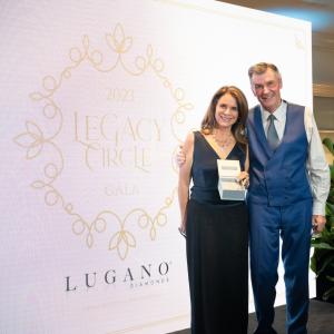 Jacqueline Dupont Carlson, Humanitarian of the Year honoree, stands on stage with Nick Jordan, Founder and CEO of Wells of Life at Legacy Circle Gala. Jacqueline holds a custom necklace designed by Lugano Diamonds.