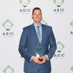 Chris is holding the AEIC trophy in his hands. He is wearing a blue suit with a light blue dress shirt and tie. He has short brown hair. Behind him is a step and repeat banner of the AEIC logo.