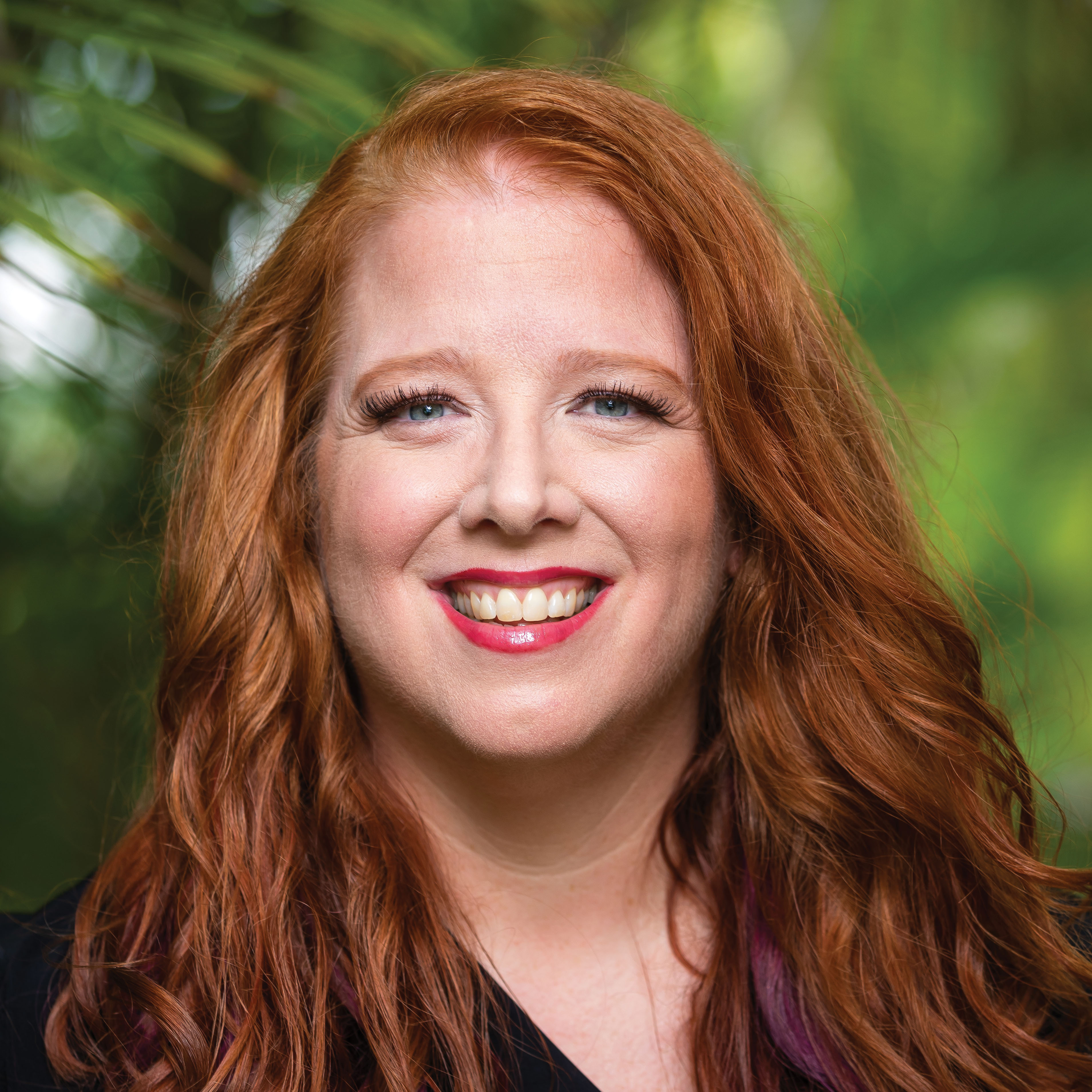 Jennifer Nash's headshot has green foliage in the background with the author wearing a black shirt. She has blue eyes and red hair.