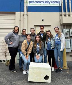 A group of volunteers stands in front of Pet Community Center. In front of them is a winter cat shelter made from a styrofoam cooler. They are wearing cat-ear headbands with the left ear clipped – the signal that a cat has been part of a trap-neuter-return program.