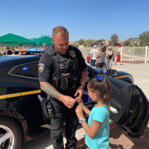 Goodyear police officer handing something to young girl standing in front of his police car