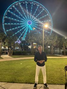 David Downing stands confidently in front of the Myrtle Beach SkyWheel, which glows with blue lights against the evening sky.
