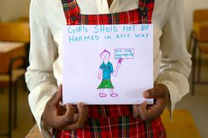 A girl in a red school uniform hold a hand drawn sign on an A4 piece of paper, which says, "Girls should not be harmed in any way - no early marriage any more"