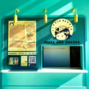 The pizza counter at Little Bettie.  Seafoam green walls with three yellow lights overhead. Large black lettering says "Pizza and Snacks''. A large yellow menu board and a round yellow sign with a drawing of "Little Bettie," a young girl wearing a slice o