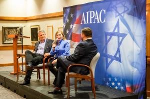 Lizzie Fletcher, wearing a blue outfit, speaks into a microphone at an AIPAC conference, flanked by two individuals, with the American and Israeli flags in the background.