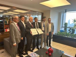 A group of five men are gathered for a formal photo opportunity in an office setting. Two of the men are holding a placard between them that reads "BLS & iDATA Partners in Growth January 2024," suggesting the celebration of a partnership or acquisition be