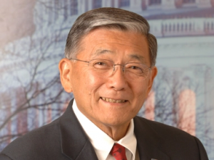 Smiling headshot of Norman Mineta wearing a black suit and red tie with the US White House in the background.