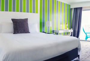 A modern room with a striking green striped wall, a plush white bed with a dark striped pillow, a sleek desk area, and a pop of color from a bright blue chair on the balcony.