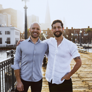 A photograph of James Rogers and Zach Gorman, the co-founders of RealReports. The two are smiling and standing side-by-side on a pier in San Francisco at sunset.