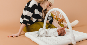 Caucasian mom engaging with baby on a white play mat with an arch with sensory toys.