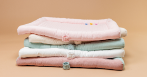 Stack of five DockATot MiniMats in each of the original colors: Blossom (pink), Sand (neutral beige), Marine (soft, neutral green/blue), Pristine White (pure white) and Multi (a combination of various chambray tones)