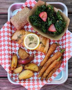 Photo of the salmon dinner served at Tanaka Farms. It is served on a metal tray with red and white checkered paper liner. The salmon is in the center of the tray and is topped with a lemon slice and fresh dill. In the bottom of the tray there are roasted 