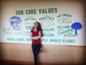 Jen Flanagan stands with hand lettered mural of Whole Foods Market core values mural in Whole Foods Market Regional Conference Room.