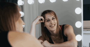 A smiling woman, Al Val leaning on a makeup table, looking at her reflection in a mirror surrounded by bright light bulbs. She has long, straight brown hair and is wearing a black tank top. Her joyful expression suggests a moment of confidence and relaxation back