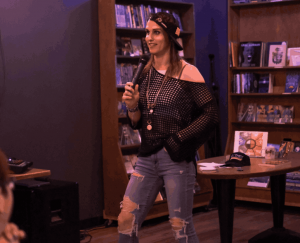 A stand-up comedian, Al Val stands in a cozy bookstore, holding a microphone with a confident smile. She's wearing a stylish off-the-shoulder mesh top, distressed jeans, and a bandana headband. The bookshelves in the background and a small table with a "T