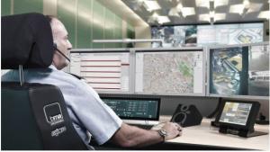 Photograph shows a male public safety officer sitting at a desk with a number of computer displays in front of him.  The image represents a modern public safety dispatch position, with the officer tasking assets in the field and monitoring radio voice com
