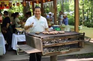 A smiling man grilling sausages on a large park grill in front of a park shelter adorned with red and yellow Spanish flags.