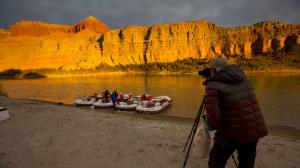 A photographer stands behind a tripod to the right of the image. The tripod is set up on a sandy beach and is pointed at four large white rafts with Holiday printed on them. The rafts are parked along the side of the river and a couple guides are on the r