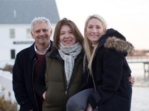 Image description A smiling family stands in front of a white building set next to water. On the right is a beautiful young woman with a warm smile and blond hair, sitting on a fence and wearing a dark parka. Her mother, wearing a scarf and standing in th