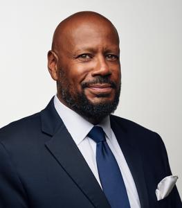A distinguished African American male with a beard, wearing a navy suit and tie, exudes confidence and professionalism against a light background.