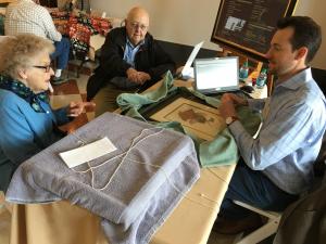 A young man, Brady Dreasher, sits at a table with a laptop and talks to an older couple who have artwork to appraise