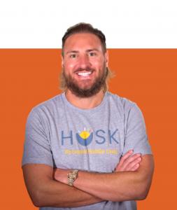 Head and shoulders image of Tony Frick, Chairman and CEO of HUSK wearing HUSK shirt. He smiles with arms crossed.