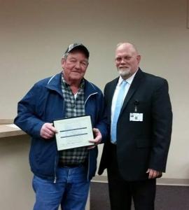 Photo of George Butts holding a certificate while stand along side Brian Costello.  George is wearing coveralls and Brian is wearing a dark suit and tie.n