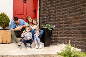 A smiling family of four is sitting in front of the red door of a house during a home swap