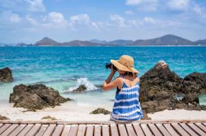 A young woman wearing a summer hat and blue and white sundress captures the breathtaking essence of a tropical paradise as she explores the sun-kissed shores on her island tour.
