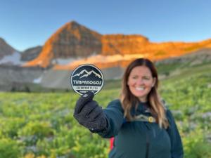 A woman holds a 2023 Timp Badge just beneath Mount Timpanogos