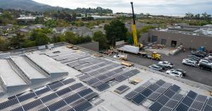 A commercial building rooftop with solar and a large crane on the other side of the. building about the lift two Tesla Megapacks onto a concrete pad.