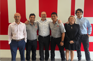Six employees of STULZ Oceania, five men and one woman, wearing company STULZ logo shirts and posing together in front of a red and white STULZ wall.